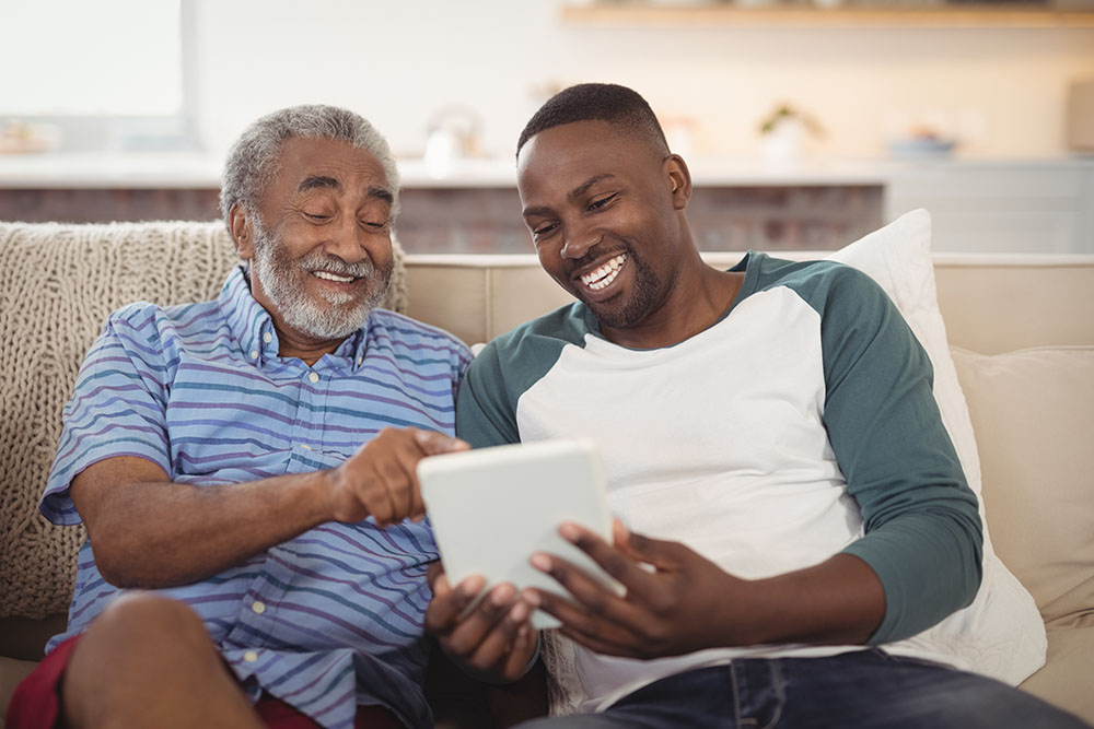 Showing how to care for an elderly parent, a son sits with his smiling father using digital tablet in living room
